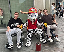 Zwei Schüler im Rollstuhl mit einem Maskottchen eines Vereins zwischen sich in der Würzburger Innenstadt.