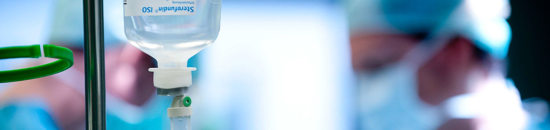 Infusion Illustrationsbild: Abbildung einer Infusion. Im Hintergrund sind arbeiten Chirurgen
