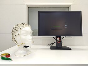 Bild vom Monitor, davor ist ein Kopfmodel mit EEG-Haube und ein Maßband 