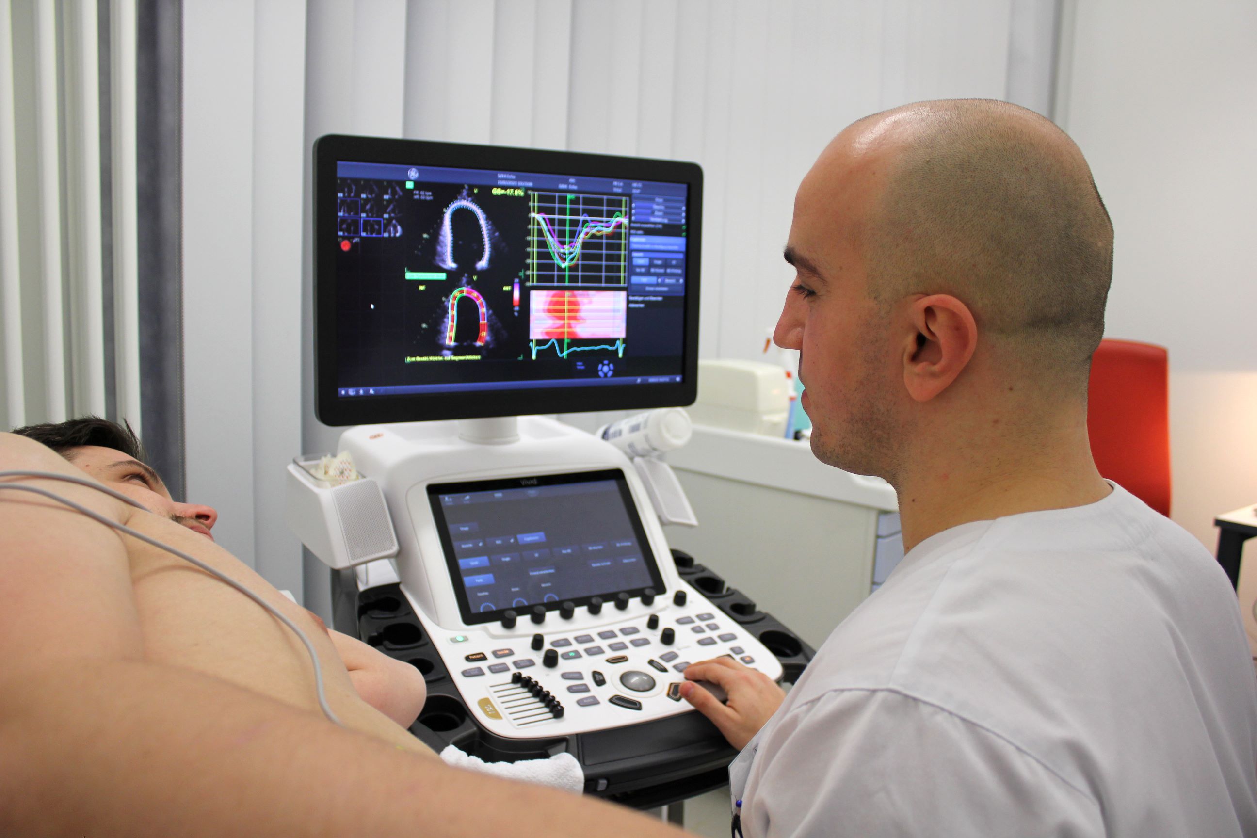 Echokardiografie Links im Anschnitt ist der Oberkörper eines liegenden Mannes zu sehen, von hinten ein Arzt, der eine Echokardiografie durchführt, in der Mitte des Bildes der Monitor mit Ultraschallbildern und Daten.