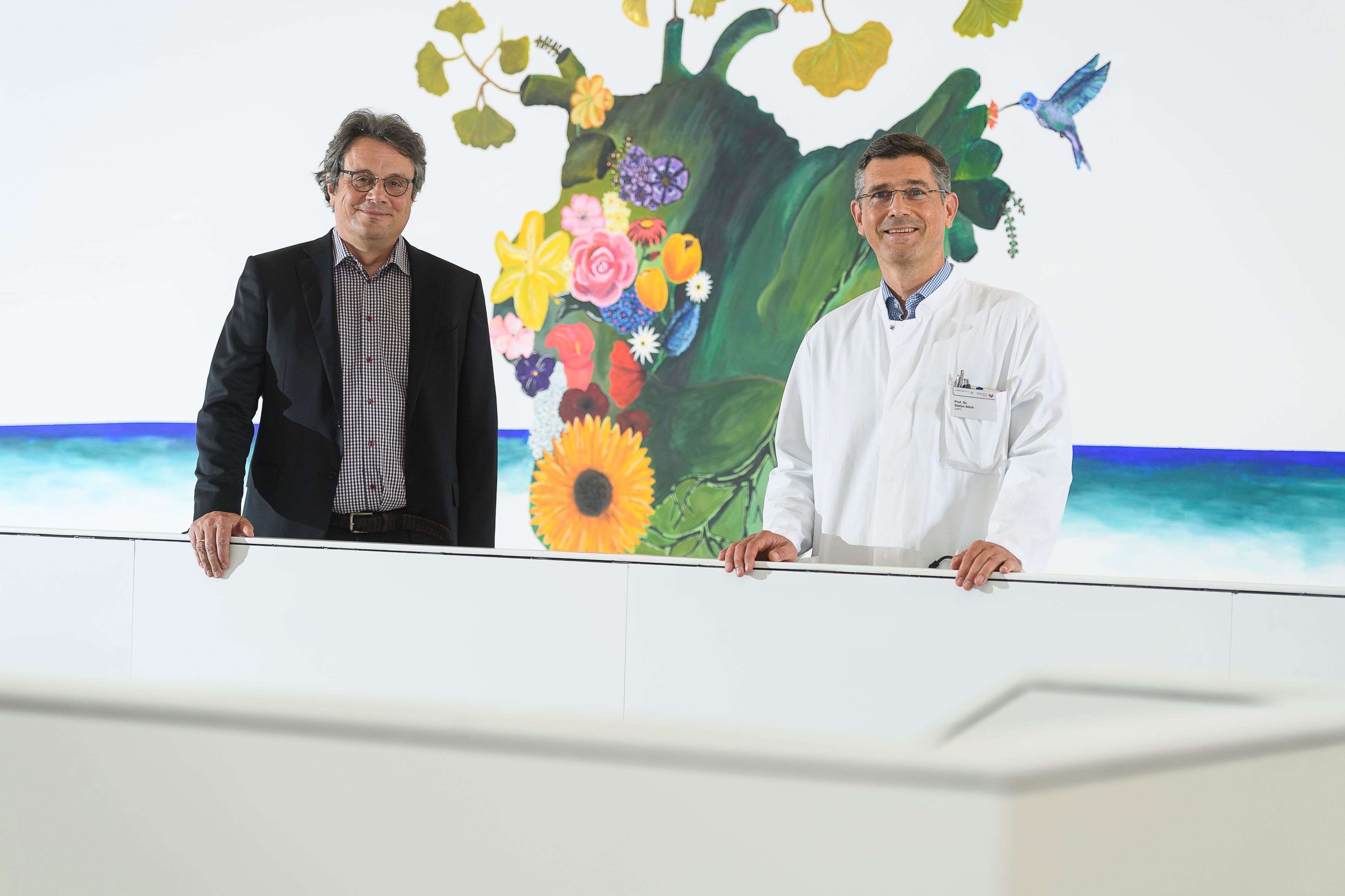 Heuschmann und Störk Peter Heuschmann in schwarzem Jacket und Stefan Störk in weißem Kittel vor einem Wandgemälde, das ein Herz darstellt, aus dem Blumen wachsen.